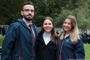 Troje studentów – uczestników Spacerku na uniwerek (dwie dziewczyny i chłopak po lewej stronie). Osoby po prawej i po lewej stronie ubrane w granatowe kurtki z białym logiem Uniwersytetu Szczecińskiego na piersi. W tle metalowy jasny płot i zielone drzewa.