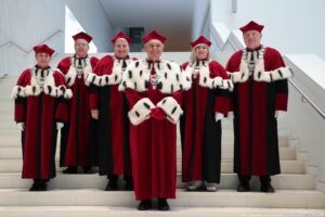 Ujęcie przodem. Wnętrze szczecińskiej filharmonii, białe tło. Na wysokich białych schodach stoi rektor Uniwersytetu Szczecińskiego (w środku) i prorektorzy (trzy kobiety, dwóch mężczyzn) ubrani w czerwone togi z gronostajami.