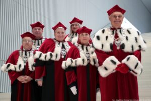 Wnętrze szczecińskiej filharmonii, białe tło. Na wysokich białych schodach stoi rektor Uniwersytetu Szczecińskiego (pierwszy z prawej) i prorektorzy (trzy kobiety, dwóch mężczyzn) ubrani w czerwone togi z gronostajami.