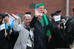 Spacerek na uniwerek. Ujęcie bokiem. Na pierwszym planie machających dwoje prodziekanów Wydziału Prawa i Administracji. Za nimi idący przedstawiciele Uniwersytetu Szczecińskiego ubrani w czarne togi. W tle ceglana ściany Bramy Królewskiej.