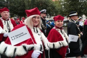 Spacerek na uniwerek. Idące w pochodzie dwie prorektorki Uniwersytetu Szczecińskiego w czerwonych togach z gronostajami. Trzymają małe białe papierowe flagi z granatowym logiem US. W tle władze innych uczelni w togach i mundurach.