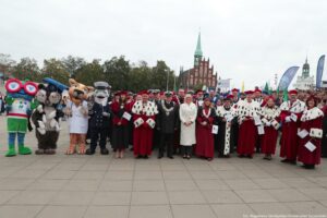 Przedstawiciele szczecińskich uczelni ubrani w uroczyste togi z gronostajami stoją razem z uczelnianymi maskotkami – sową USZatką w birecie, PUMą w kitlu, Morświnem w marynarskim mundurze i robotem. W tle kościół św. św. Piotra i Pawła oraz Zamek Książąt Pomorskich.