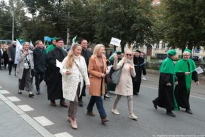 Spacerek na uniwerek. Ujęcie bokiem. Idący przedstawiciele Uniwersytetu Szczecińskiego ubrani w czarne togi z zielonymi elementami, trzymający białe papierowe flagi. W tle drzewa i budynki.