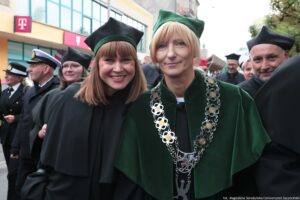 Spacerek na uniwerek. Ujęcie przodem. Idący przedstawiciele Uniwersytetu Szczecińskiego ubrani w czarne togi z zielonymi elementami, trzymający. Na pierwszym planie dwie uśmiechnięte kobiety.