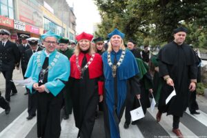 Spacerek na uniwerek. Ujęcie przodem. Idący przedstawiciele Uniwersytetu Szczecińskiego ubrani w czarne togi z zielonymi, czerwonymi i niebieskimi elementami, trzymający białe papierowe flagi. W tle inni uczestnicy wydarzenia, drzewa i budynki.