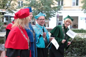 Spacerek na uniwerek. Ujęcie bokiem. Idący przedstawiciele Uniwersytetu Szczecińskiego ubrani w czarne togi z zielonymi, czerwonymi i niebieskimi elementami, trzymający białe papierowe flagi. W tle drzewa i budynki.