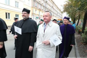 Spacerek na uniwerek. Ujęcie na kanclerza Uniwersytetu Szczecińskiego trzymającego małą białą papierową flagę z grantowym logiem uczelni. Obok mężczyzna w czarnej todze. W tle uczestnicy wydarzenia w togach z elementami w różnych kolorach.