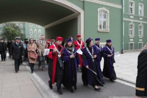 Spacerek na uniwerek. Idący przedstawiciele szczecińskich uczelni ubrani w uroczyste togi mijają zielony gmach Urzędu Miejskiego w Szczecinie.