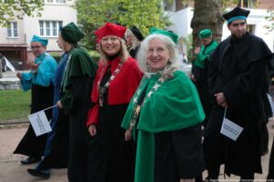 Spacerek na uniwerek. Ujęcie bokiem. Idący przedstawiciele Uniwersytetu Szczecińskiego ubrani w czarne togi z zielonymi, czerwonymi i niebieskimi elementami, trzymający białe papierowe flagi. W tle drzewa i budynki.