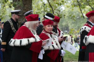 Spacerek na uniwerek. Park. Idące w pochodzie dwie prorektorki Uniwersytetu Szczecińskiego w czerwonych togach z gronostajami. Trzymają małe białe papierowe flagi z granatowym logiem US. W tle władze innych uczelni w togach i mundurach.