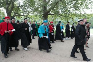 Spacerek na uniwerek. Ujęcie bokiem. Idący przedstawiciele szczecińskich uczelni ubrani w uroczyste togi i trzymający białe papierowe flagi. W tle drzewa.