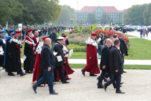 Spacerek na uniwerek. Ujęcie bokiem. Idący oficjele i przedstawiciele szczecińskich uczelni ubrani w uroczyste togi z gronostajami i trzymający białe papierowe flagi. W tle gmach Urzędu Miejskiego w Szczecinie i inni uczestnicy pochodu.