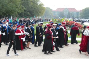 Spacerek na uniwerek. Ujęcie bokiem. Idący przedstawiciele szczecińskich uczelni ubrani w uroczyste togi z gronostajami i trzymający białe papierowe flagi. W tle gmach Urzędu Miejskiego w Szczecinie i inni uczestnicy pochodu.