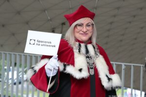 Jasne tło. Ujęcie do pasa. Na zdjęciu uśmiechnięta prorektor Uniwersytetu Szczecińskiego w czerwonej todze z gronostajami. Trzyma małą białą papierową flagę z logiem Spacerku na uniwerek.