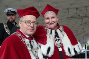 Jasne tło. Ujęcie do piersi. Na zdjęciu prorektorzy Uniwersytetu Szczecińskiego w czerwonych togach z gronostajami. W tle mężczyzna w mundurze.