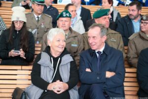 Inauguracja roku akademickiego. Widownia. Ujęcie na dwoje oficjeli z przodu (kobieta i mężczyzna). W tle przedstawiciele Wojska Polskiego w mundurach i inni uczestnicy wydarzenia.