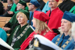 Inauguracja roku akademickiego. Widownia. Ujęcie na sześcioro przedstawicieli Uniwersytetu Szczecińskiego w czarnych togach z zielonymi, granatowymi i czerwonymi elementami. W tle inni uczestnicy wydarzenia.