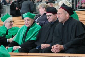 Inauguracja roku akademickiego. Widownia. Ujęcie na czworo przedstawicieli Uniwersytetu Szczecińskiego w czarnych togach z zielonymi i bordowymi elementami. W tle inni uczestnicy wydarzenia.