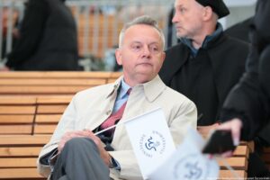 Inauguracja roku akademickiego. Widownia. Ujęcie na kanclerza Uniwersytetu Szczecińskiego trzymającego małą białą papierową flagę z grantowym logiem uczelni. W tle po prawej mężczyzna w czarnej todze.