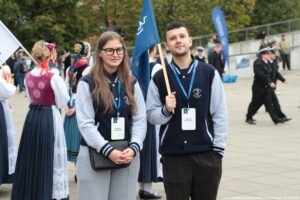 Spacerek na uniwerek. Dwoje studentów w bluzach z logiem Uniwersytetu Szczecińskiego. Chłopak trzyma granatową flagę. W tle inni uczestnicy wydarzenia.