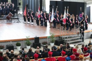 Inauguracja roku akademickiego. Widownia (ujęcie tyłem). Widok na scenę. Po lewej ubrany na czarno chór, po prawej poczty sztandarowe.