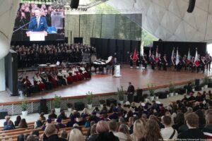 Inauguracja roku akademickiego. Widownia (ujęcie tyłem). Widok na scenę. Po lewej rektorzy szczecińskich uczelni w uroczystych togach, na środku ubrany na czarno chór, po prawej poczty sztandarowe. Na środku za białą mównicą przedstawiciel Kancelarii Prezydenta RP. U góry ekran z przemówieniem.