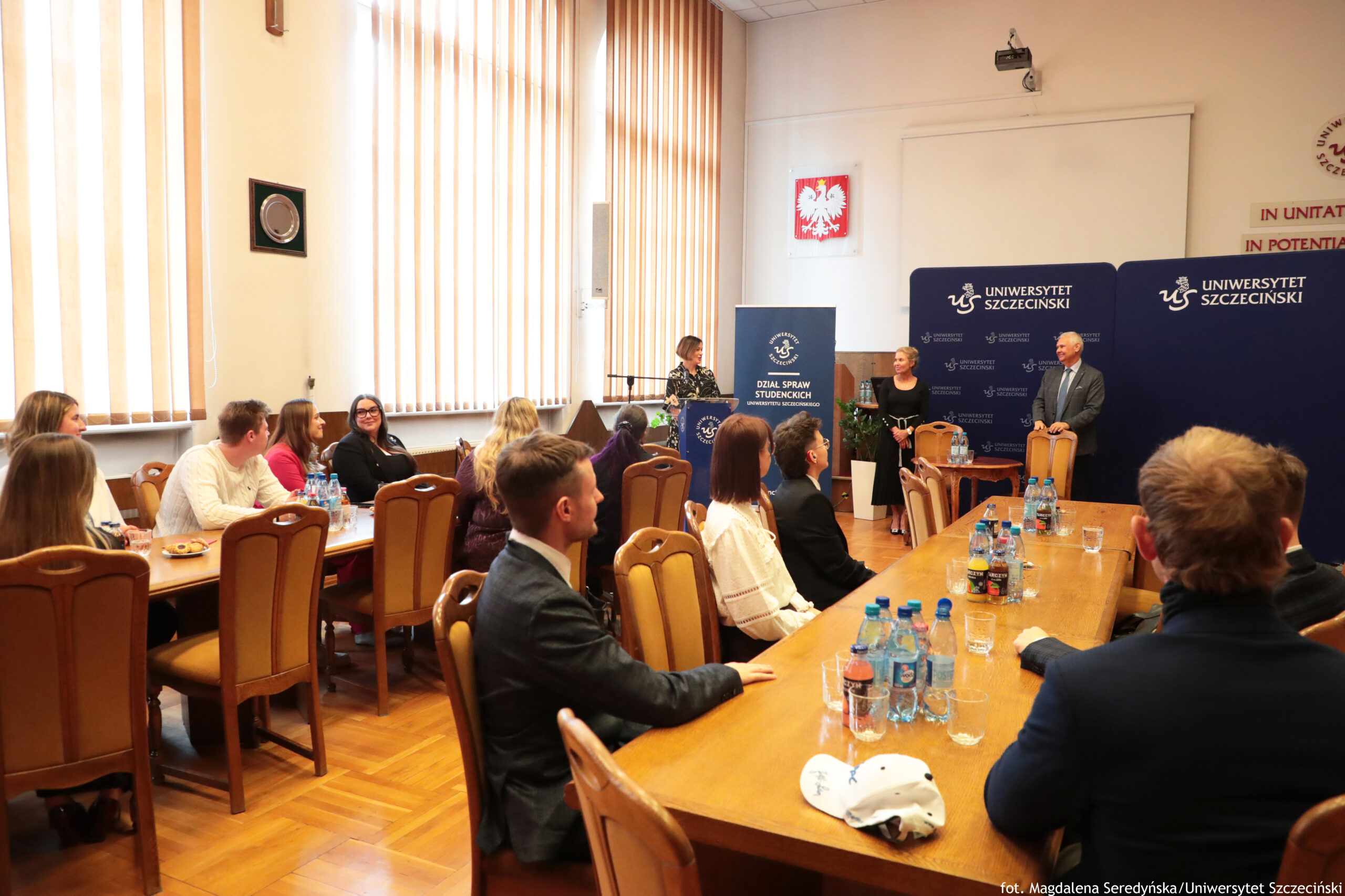 Sala Senatu US z drewnianymi stołami i krzesłami wypełniona osobami uczestniczącymi w spotkaniu. W centrum i po prawej stronie ludzie siedzą przy długich stołach, zwróceni w stronę przodu sali, gdzie stoją trzy osoby przy niebieskich ściankach z logo i napisem „Uniwersytet Szczeciński”. Jedna kobieta stoi za mównicą z napisem „Dział Spraw Studenckich”, druga kobieta i mężczyzna w garniturze stoją obok niej, jakby prowadzili lub otwierali wydarzenie. Na stołach stoją głównie butelki z wodą i napojami, są też szklanki oraz na jednym z bliższych stołów jasna czapka z daszkiem. Po lewej stronie przy innym stole siedzi kilka osób rozmawiających między sobą, w tle widać wysokie okna z pionowymi żaluzjami i na ścianie biało‑czerwony godło Polski.