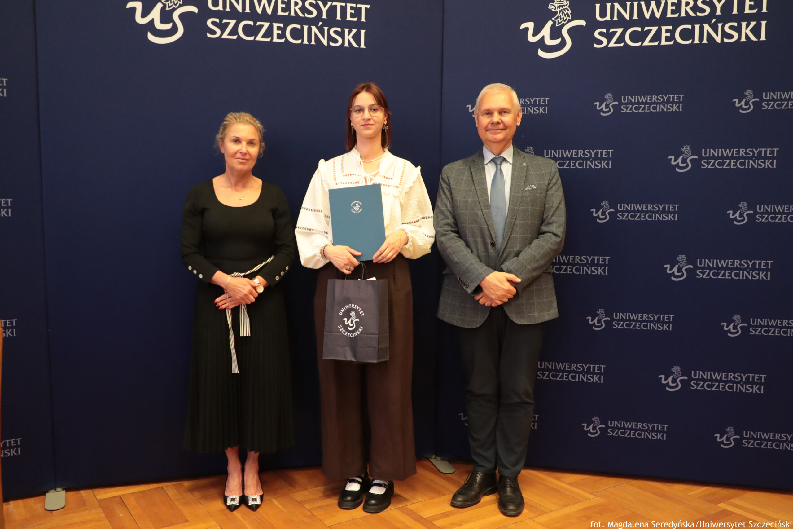 Trzy osoby stojące na tle granatowej ścianki z napisem „Uniwersytet Szczeciński”. W centrum stoi młoda osoba o jasnej skórze, w okularach, z włosami do ramion. Ubrana jest w białą, szeroką bluzkę z ażurowymi zdobieniami i brązowe, szerokie spodnie. Na nogach ma czarno‑białe buty. Trzyma granatową teczkę z białym logo uczelni oraz małą granatową torbę z tym samym logo. Po lewej stronie stoi kobieta o jasnej skórze, z upiętymi włosami. Ma na sobie długą, czarną sukienkę z długimi rękawami i srebrzysty pasek zawiązany z przodu. Nosi czarne buty na obcasie z ozdobną klamrą. Po prawej stronie stoi mężczyzna o jasnej skórze, z krótkimi siwymi włosami. Ma na sobie szarą, kratowaną marynarkę, błękitną koszulę, jasnoniebieski krawat i ciemne spodnie. Nosi ciemne skórzane buty. Tło stanowi jednolita granatowa plansza z wielokrotnie powtórzonym białym napisem „UNIWERSYTET SZCZECIŃSKI” oraz logo uczelni. Podłoga jest drewniana, w ciepłym, jasnobrązowym kolorze.