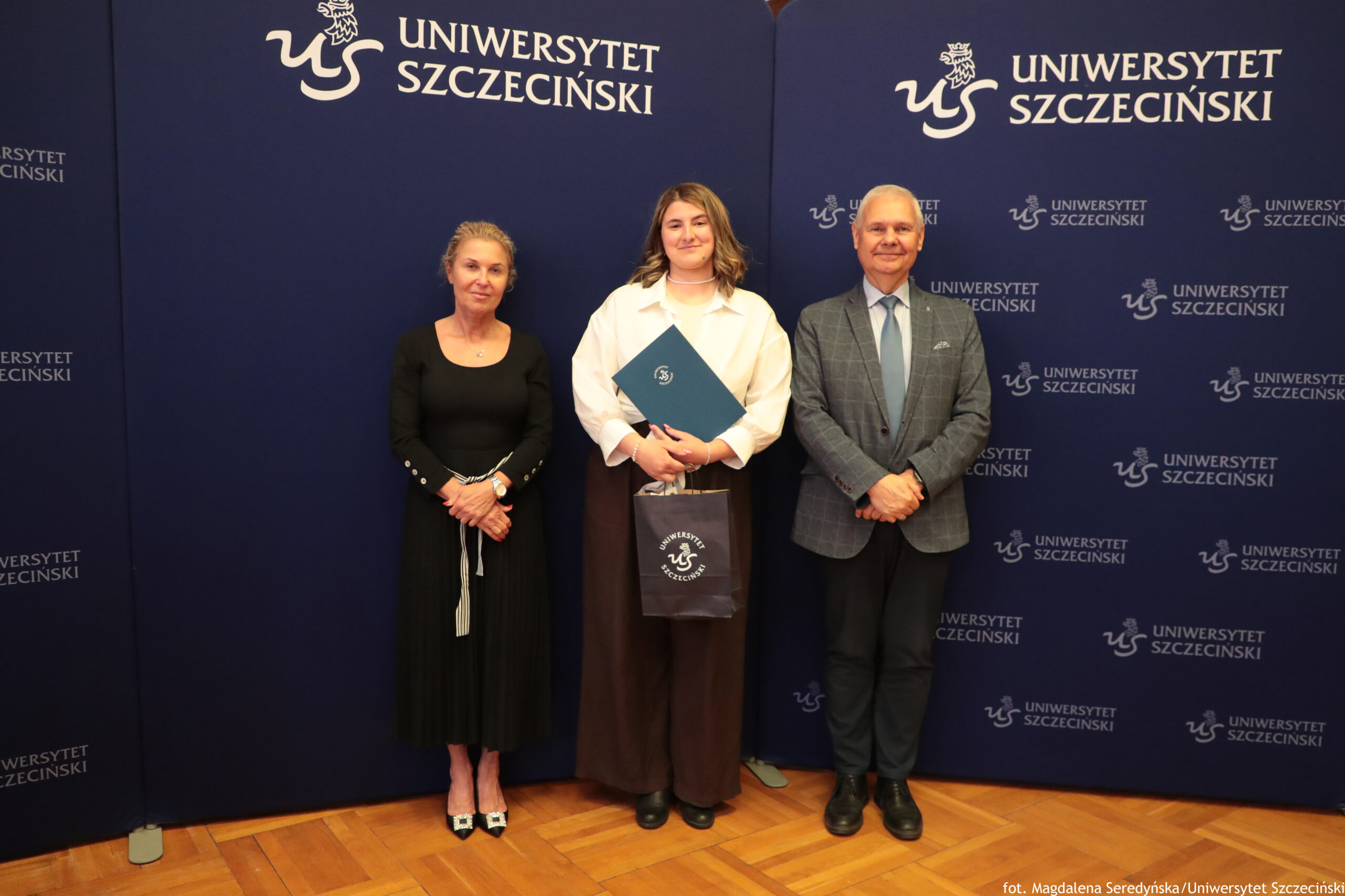 Trzy osoby stojące na tle granatowej ścianki z logo i napisem „Uniwersytet Szczeciński”. Po lewej stoi kobieta o jasnej karnacji i upiętych, siwiejących włosach, ubrana w długą, czarną sukienkę z długimi rękawami; ma na ręku zegarek i jasne buty na obcasie z ozdobną klamrą. W środku stoi młoda osoba o jasnej karnacji i średniej długości, falowanych, jasnobrązowych włosach, ubrana w luźną białą koszulę i szerokie, ciemne spodnie; trzyma granatową teczkę oraz granatową torbę z okrągłym logo Uniwersytetu Szczecińskiego. Po prawej stoi mężczyzna o jasnej karnacji i krótkich, siwych włosach, ubrany w błękitną koszulę, jasnoniebieski krawat, szarą marynarkę w kratę i ciemne spodnie; ma czarne buty. Tło stanowią granatowe panele z wielokrotnie powtórzonym białym napisem „UNIWERSYTET SZCZECIŃSKI” oraz logo uczelni.