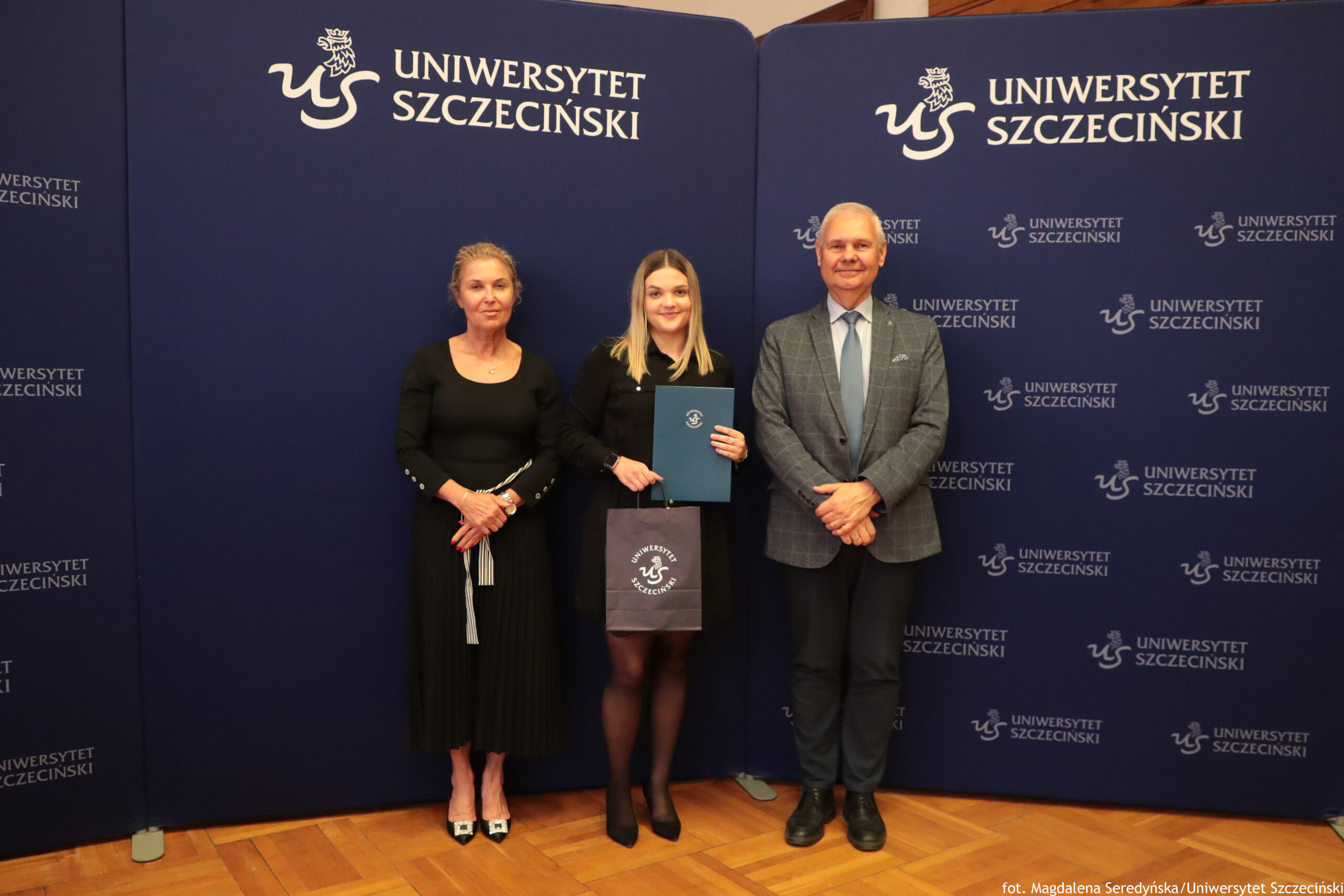Trzy osoby stojące na tle ścianki z logo Uniwersytetu Szczecińskiego. W centrum znajduje się młoda kobieta o jasnej cerze i prostych blond włosach do ramion, ubrana na czarno. Trzyma granatową teczkę z białym emblematem uczelni oraz granatową torebkę z tym samym logo. Po jej lewej stronie stoi kobieta o jasnej cerze, w długiej czarnej sukience z długimi rękawami, z upiętymi włosami w kolorze blond. Ma założone ręce, na nogach jasne pantofle na obcasie. Po prawej stronie stoi mężczyzna o jasnej cerze, w szarej kraciastej marynarce, jasnoniebieskiej koszuli i dopasowanym krawacie, w ciemnych spodniach i czarnych butach. Tło stanowi granatowa ścianka z wielokrotnie powtórzonym białym napisem „UNIWERSYTET SZCZECIŃSKI” i logo uczelni. Podłoga jest z jasnego parkietu.