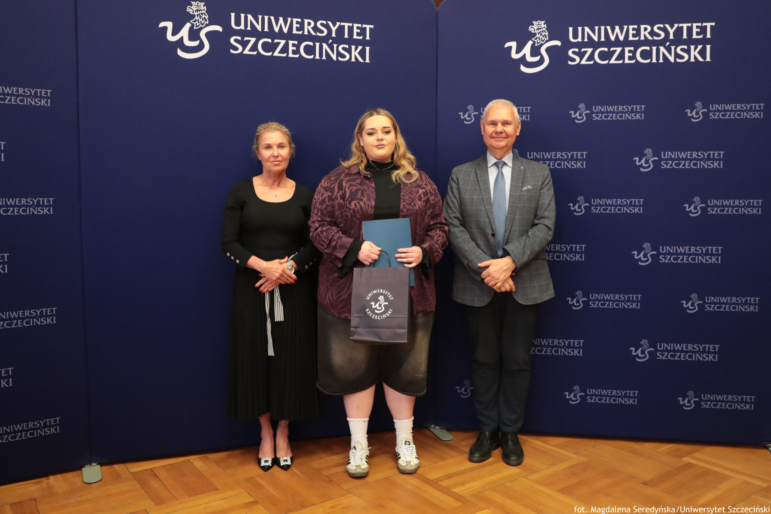 Trzy osoby stojące na tle ścianki z napisem „Uniwersytet Szczeciński”. Po lewej stronie stoi kobieta o jasnej skórze, z upiętymi z tyłu blond włosami, ubrana w długą, czarną sukienkę do połowy łydki i czarne buty na obcasie z jasnymi czubkami; na rękach ma zegarek i bransoletkę, dłonie złożone przed sobą. Na środku stoi młoda osoba o jasnej skórze i długich, falowanych, blond włosach. Ma na sobie czarną koszulkę, luźną bordowo‑czarną kurtkę z wzorem, ciemne krótkie spodnie za kolano, białe skarpetki i białe tenisówki. Trzyma ciemną, prostokątną teczkę oraz granatową torbę z białym logo i napisem „Uniwersytet Szczeciński”. Po prawej stronie stoi mężczyzna o jasnej skórze, siwych, krótkich włosach, ubrany w jasnoniebieską koszulę, niebieski krawat, szarą marynarkę w kratę i ciemne spodnie z czarnymi butami; ręce ma złożone z przodu. Tło stanowi granatowa ścianka z wielokrotnie powtórzonym białym logo i napisem „UNIWERSYTET SZCZECIŃSKI”. Podłoga jest z jasnego, drewnianego parkietu.