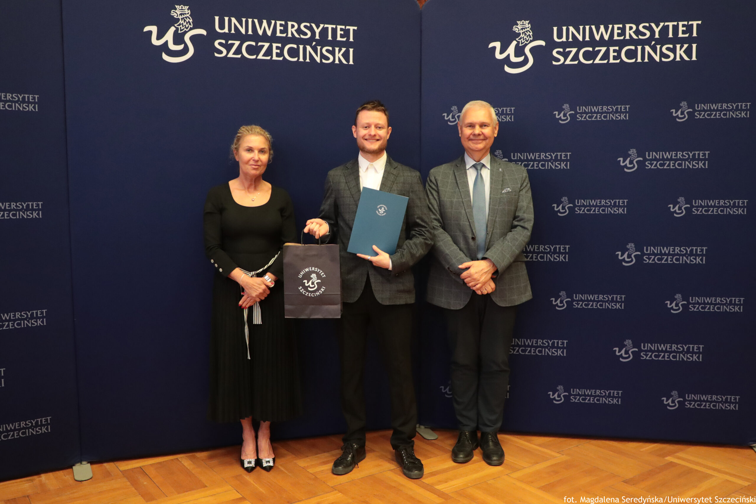 Trzy osoby stoją na tle granatowych ścian z białym napisem „Uniwersytet Szczeciński” i logotypem uczelni. Po lewej stronie stoi kobieta o jasnej skórze i związanych włosach, ubrana w długą czarną sukienkę z długimi rękawami i czarne lakierowane buty na obcasie; trzyma ręce przed sobą. W środku stoi młody mężczyzna o jasnej skórze w szarej kraciastej marynarce, białej koszuli i ciemnych spodniach; uśmiecha się, w lewej ręce trzyma niebieską teczkę z herbem Uniwersytetu Szczecińskiego, w prawej ciemną torbę prezentową z tym samym logo. Po prawej stronie stoi starszy mężczyzna o jasnej skórze w podobnej szarej kraciastej marynarce, błękitnej koszuli, jasnym krawacie i ciemnych spodniach; ma skrzyżowane dłonie przed sobą i również się uśmiecha. Podłoga jest z jasnego parkietu. W prawym dolnym rogu małym białym tekstem widoczny jest podpis: „fot. Magdalena Seredyńska/Uniwersytet Szczeciński”.