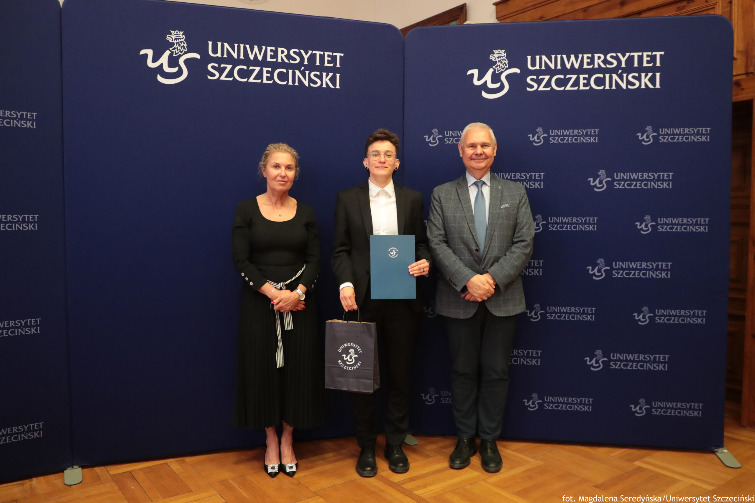 Trzy osoby stoją na tle granatowych ścian z białym napisem „Uniwersytet Szczeciński” i logotypem uczelni. Po lewej stronie stoi kobieta o jasnej skórze i związanych włosach, ubrana w długą czarną sukienkę z długimi rękawami i czarne lakierowane buty na obcasie; trzyma ręce przed sobą. W środku stoi młody mężczyzna o jasnej skórze w szarej kraciastej marynarce, białej koszuli i ciemnych spodniach; uśmiecha się, w lewej ręce trzyma niebieską teczkę z herbem Uniwersytetu Szczecińskiego, w prawej ciemną torbę prezentową z tym samym logo. Po prawej stronie stoi starszy mężczyzna o jasnej skórze w podobnej szarej kraciastej marynarce, błękitnej koszuli, jasnym krawacie i ciemnych spodniach; ma skrzyżowane dłonie przed sobą i również się uśmiecha. Podłoga jest z jasnego parkietu. W prawym dolnym rogu małym białym tekstem widoczny jest podpis: „fot. Magdalena Seredyńska/Uniwersytet Szczeciński”.