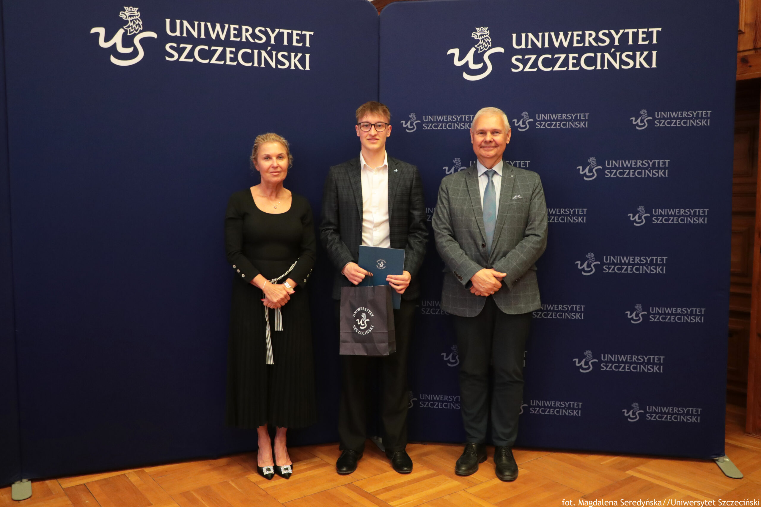 Trzy osoby stojące na tle granatowej ścianki z napisem „Uniwersytet Szczeciński”. Po lewej stronie stoi kobieta w czarnej, długiej sukience z długim rękawem, z jasnymi włosami upiętymi do tyłu i w jasnych butach na obcasie. Na środku stoi młody mężczyzna w okularach, białej koszuli i ciemnej marynarce w kratę; trzyma granatową teczkę oraz granatową torbę z białym logo Uniwersytetu Szczecińskiego. Po prawej stronie stoi starszy mężczyzna w jasnoniebieskiej koszuli, z krawatem i w szarej marynarce w kratę, z rękami złożonymi przed sobą. Za nimi znajduje się granatowe tło z wielokrotnie powtórzonym białym logo i napisem „UNIWERSYTET SZCZECIŃSKI”; podłoga jest drewniana, w ciepłym kolorze.