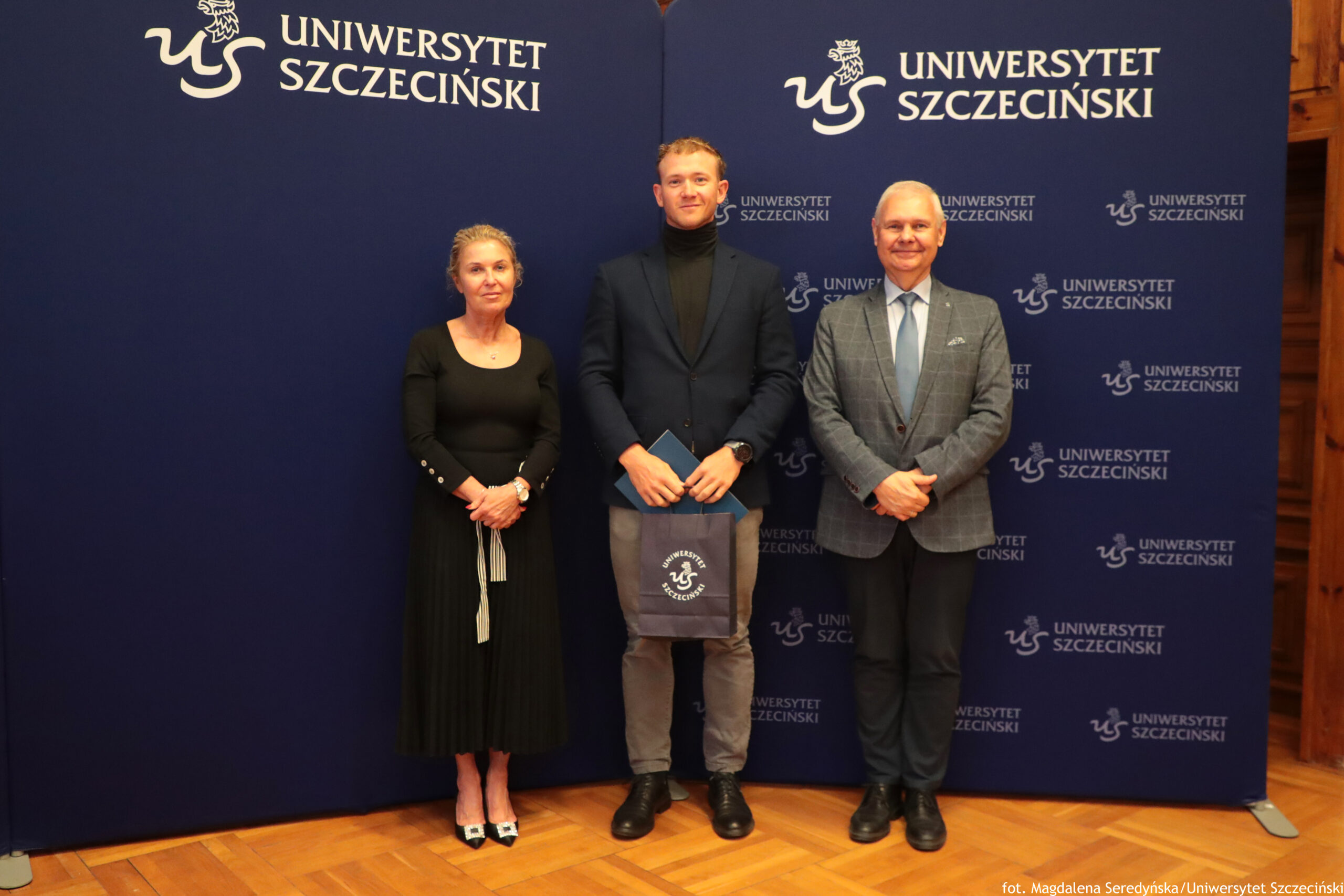 Trzy osoby stojące na tle ściany z logotypem Uniwersytetu Szczecińskiego. W środku stoi młody mężczyzna o jasnych włosach, ubrany w ciemny golf, granatową marynarkę i szare spodnie. W rękach trzyma granatową teczkę i szarą torbę z białym logo Uniwersytetu Szczecińskiego. Po jego lewej stronie znajduje się starsza kobieta o jasnych, upiętych włosach, ubrana w długą, czarną sukienkę z długimi rękawami oraz czarne buty na obcasie ze srebrnymi klamrami. Po prawej stronie mężczyzny stoi starszy mężczyzna o siwych włosach, ubrany w jasnoniebieską koszulę, błękitny krawat, szarą marynarkę w kratę i ciemne spodnie; ma na nogach czarne buty. Tło stanowią dwa granatowe panele z białymi napisami „UNIWERSYTET SZCZECIŃSKI” oraz powtarzającym się logo uczelni. Podłoga jest drewniana, w ciepłym odcieniu.