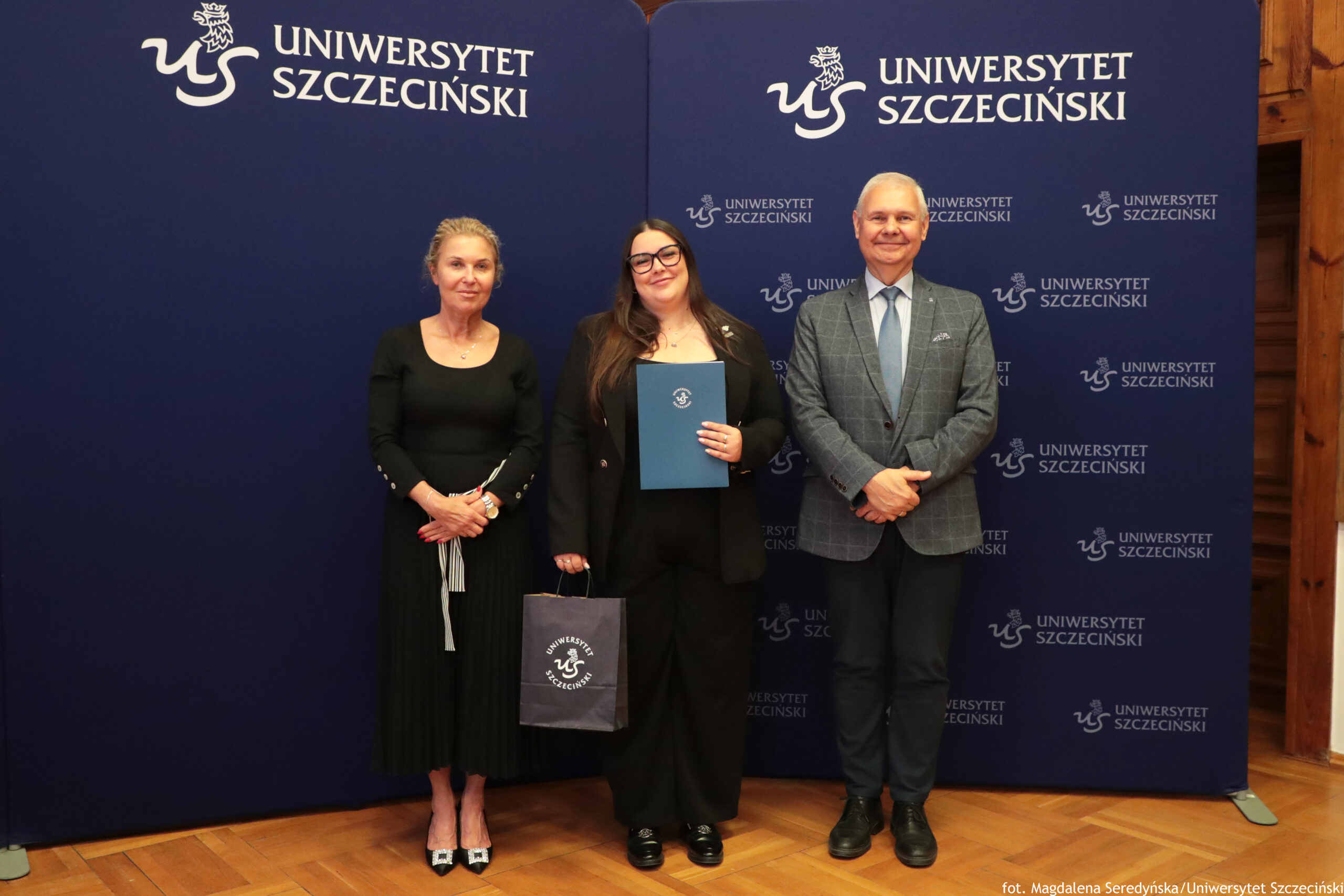 Trzy osoby stojące na tle granatowej ścianki z logotypem Uniwersytetu Szczecińskiego. W tle widać jednolitą granatową planszę z białym napisem „Uniwersytet Szczeciński” i skrótem „US” powtarzającym się w kilku miejscach. Po lewej stronie stoi kobieta o jasnej skórze i jasnych, upiętych włosach. Ma na sobie dopasowaną, czarną sukienkę do połowy łydki, z plisowanym dołem i jasną, pionową wstążką przy talii. Na stopach ma czarne buty na obcasie z jasnymi czubkami. Na nadgarstku ma zegarek, na szyi delikatny naszyjnik. Trzyma dłonie splecione przed sobą. W środku stoi kobieta o jasnej skórze i długich, ciemnych włosach opadających na ramiona. Nosi czarny garnitur: długą marynarkę i czarne szerokie spodnie, pod spodem ciemny top. Na nosie ma ciemne okulary w grubych oprawkach. W jednej ręce trzyma granatową teczkę lub dyplom z herbem Uniwersytetu Szczecińskiego, w drugiej granatową torbę papierową z tym samym logo. Na klapie marynarki przypięta jest mała przypinka. Po prawej stronie stoi mężczyzna o jasnej skórze i krótkich, siwych włosach. Ma na sobie szarą marynarkę w kratę, jasnoniebieską koszulę, jasny krawat i ciemne spodnie. Na nogach ma ciemne buty. Stoi z dłońmi złożonymi z przodu i uśmiecha się. Podłoga jest z jasnego, drewnianego parkietu. W prawym dolnym rogu, przy krawędzi zdjęcia, znajdują się małe białe napisy z informacją o autorce zdjęcia i uczelni.