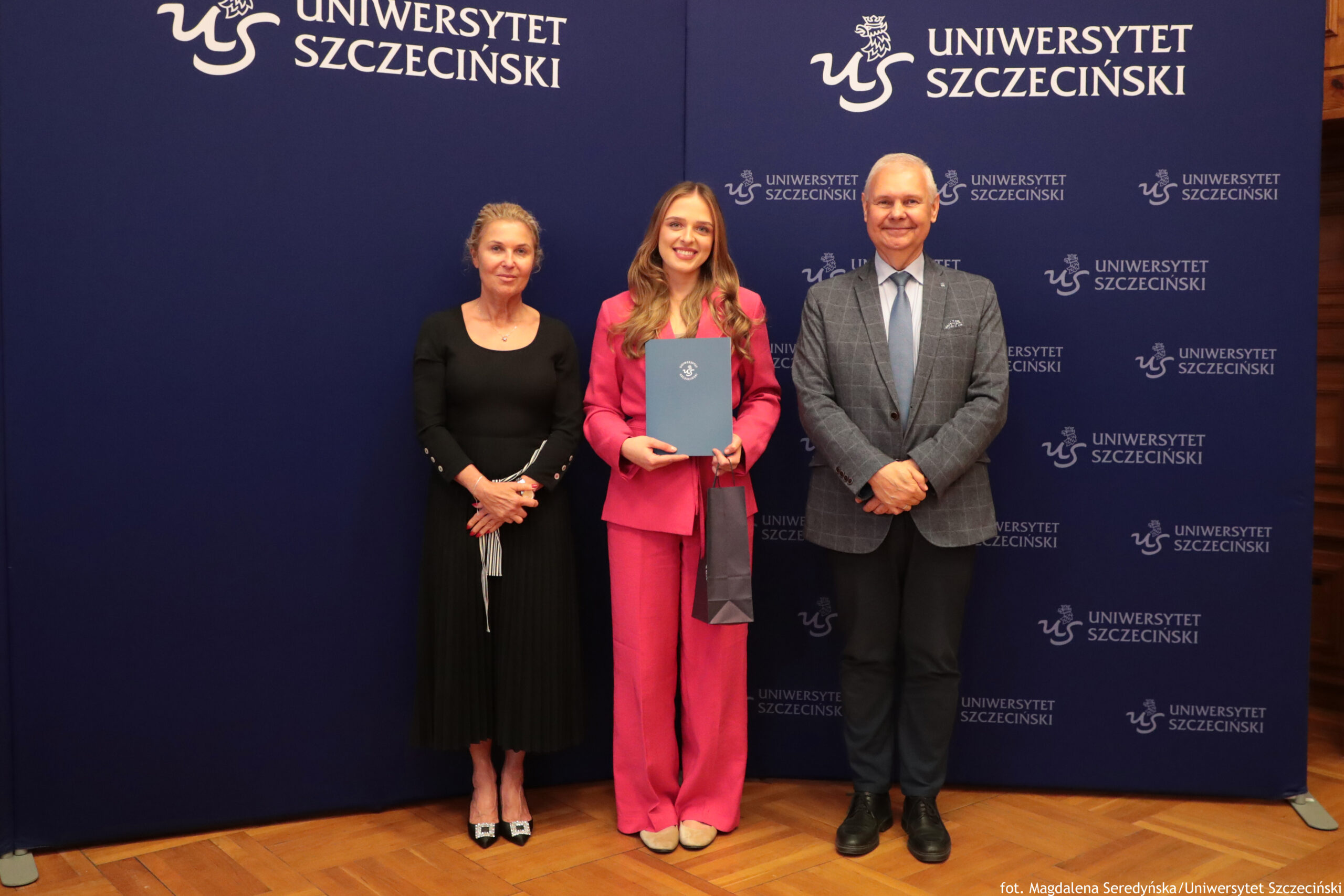 Trzy osoby stoją na tle granatowej ścianki z logo i napisem „Uniwersytet Szczeciński”. Po lewej stronie stoi kobieta o jasnych włosach upiętych do tyłu, o jasnej karnacji. Ma na sobie dopasowaną, czarną sukienkę o długości za kolano, z długimi rękawami i rozkloszowanym dołem z plisami. Na stopach ma czarne buty na obcasie z jasną ozdobą z przodu. Ręce trzyma splecione przed sobą, w dłoniach fragment jasnego materiału, który zwisa pionowo. W środku stoi młoda kobieta o jasnej karnacji i długich, rozpuszczonych, falowanych włosach w odcieniu ciemny blond. Ubrana jest w jaskraworóżowy garnitur: marynarkę i szerokie spodnie. Pod marynarką ma jasną bluzkę. W obu dłoniach trzyma niebieską teczkę z białym okrągłym logo Uniwersytetu Szczecińskiego, skierowaną frontem do przodu. Obok teczki zwisa w dół podłużna, ciemna torebka prezentowa. Na nogach ma jasne buty. Uśmiecha się. Po prawej stronie stoi mężczyzna o jasnej karnacji i krótkich siwych włosach. Ma na sobie jasnoniebieską koszulę, szaroniebieski krawat i szarą marynarkę w kratę. Do tego nosi ciemne spodnie i czarne buty. Ręce ma skrzyżowane z przodu na wysokości brzucha, na twarzy ma uprzejmy uśmiech. Tło stanowią dwa połączone, granatowe panele. Po lewej stronie, w górnej części, widoczne są białe napisy: „US” – stylizowane logo – oraz „UNIWERSYTET SZCZECIŃSKI”. Po prawej stronie, w górze, powtórzone są logo i ten sam napis. Pośrodku oraz po prawej stronie, niżej, kilka razy powtarza się mniejsze logo z napisem „UNIWERSYTET SZCZECIŃSKI”. Na dole, przy prawym dolnym rogu, małym białym tekstem podano informację: „fot. Magdalena Seredyńska/Uniwersytet Szczeciński”. Pod stopami osób widoczna jest drewniana podłoga z ułożonymi w jodełkę panelami.