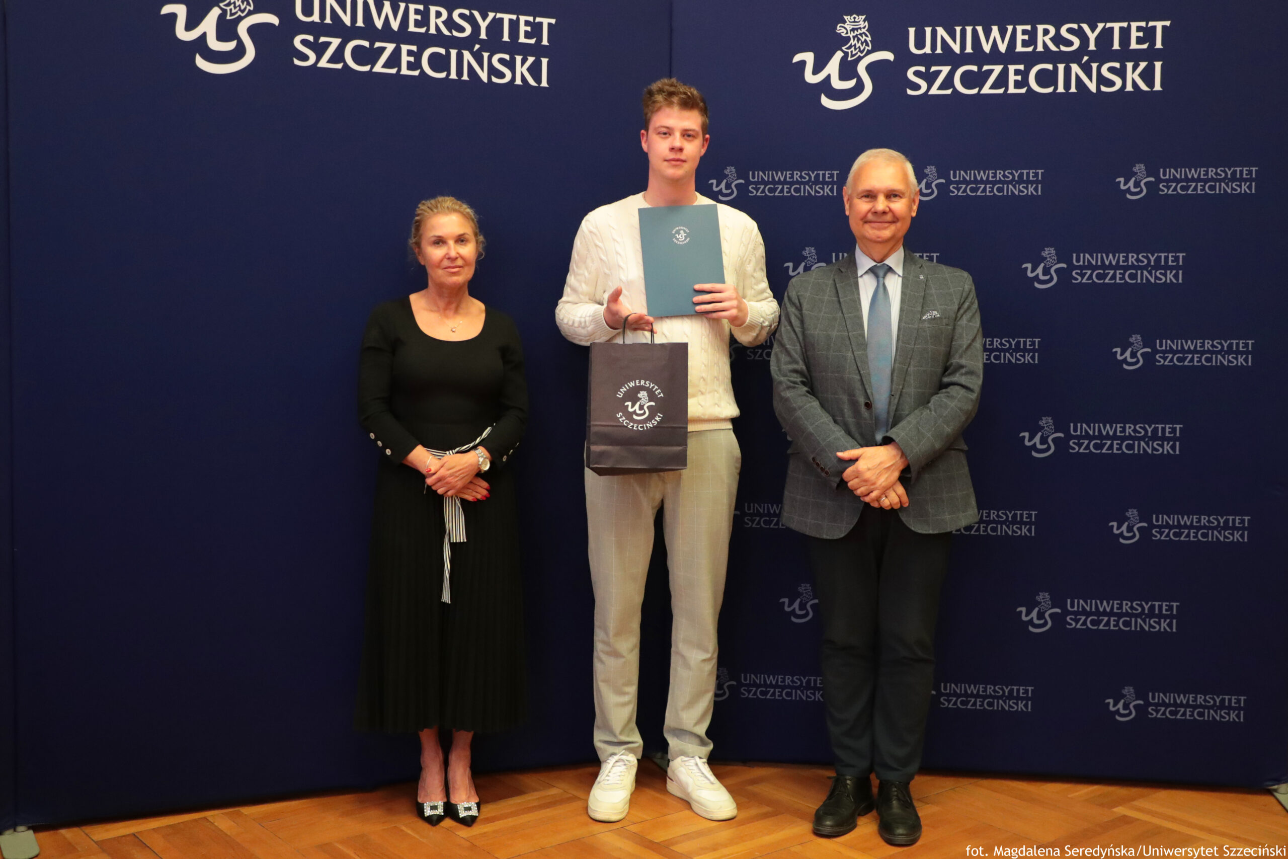Trzy osoby stojące na tle granatowej ścianki z napisem „Uniwersytet Szczeciński”. Po lewej kobieta o jasnych, upiętych włosach, jasnej karnacji, w czarnej, długiej sukni z długim rękawem i w butach na obcasie z jasnymi czubkami; dłonie złożone przed sobą. W środku młody mężczyzna o jasnej karnacji, w kremowym swetrze z wzorem warkoczowym, jasnych materiałowych spodniach i białych sportowych butach; trzyma przed sobą niebieską teczkę z białym logo uczelni oraz granatową papierową torbę z tym samym logo. Po prawej mężczyzna o jasnej karnacji, siwych włosach, w szarej kraciastej marynarce, jasnoniebieskiej koszuli, jasnym krawacie, ciemnych spodniach i ciemnych butach; ręce złożone przed sobą. Tło stanowi jednolita granatowa ścianka z wielokrotnie powtórzonym białym logo „US” i napisem „UNIWERSYTET SZCZECIŃSKI”. Na dole po prawej drobnym drukiem podpis zdjęcia: „fot. Magdalena Seredyńska/Uniwersytet Szczeciński”.