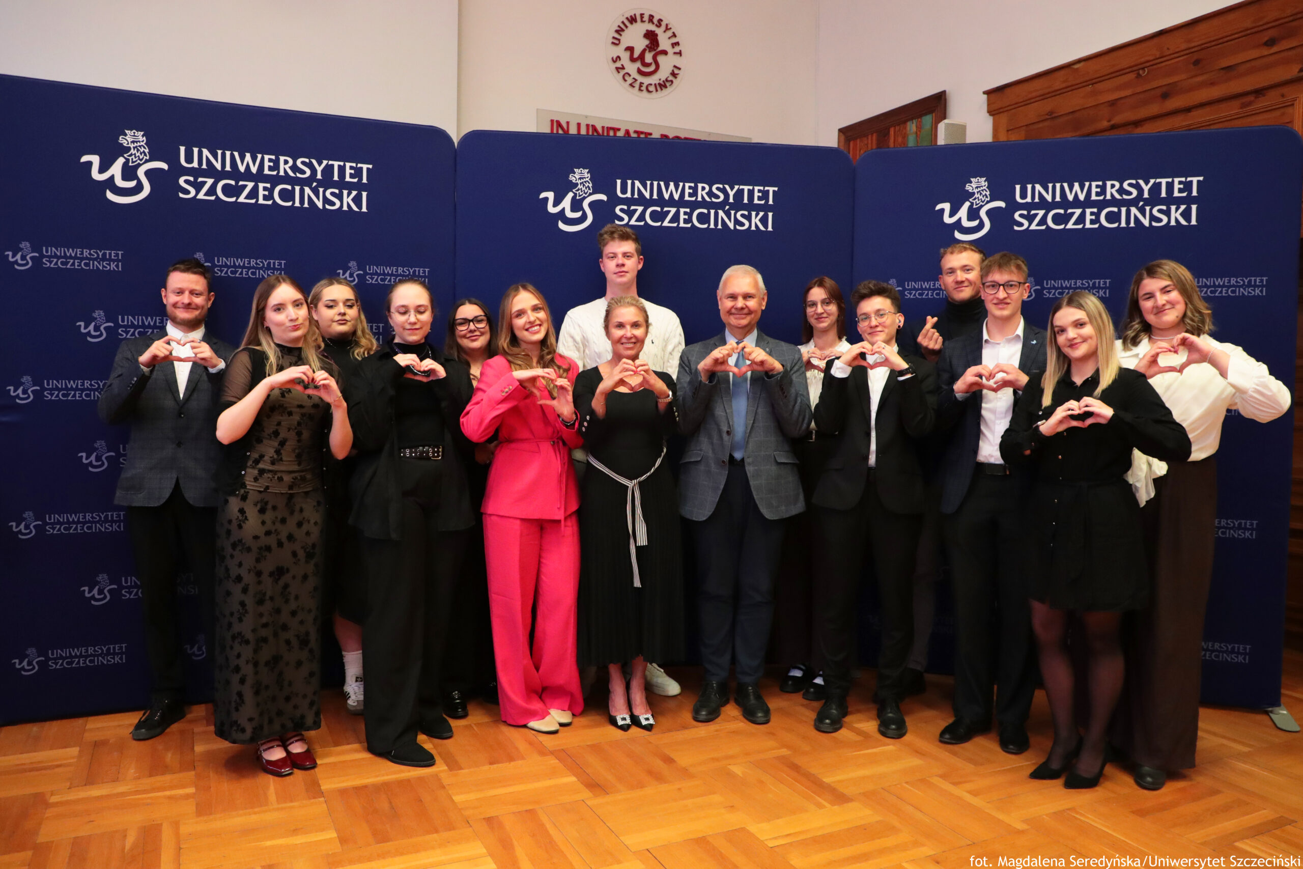 Grupa trzynastu osób stojących na tle ściany z logo „Uniwersytet Szczeciński”. Wszyscy stoją w jednym rzędzie na parkiecie z jasnego drewna i trzymają dłonie przed sobą ułożone w kształt serca. W tle znajdują się trzy granatowe ścianki z białym napisem „Uniwersytet Szczeciński” i logo uczelni po lewej stronie tekstu; napis powtarza się wielokrotnie. Osoby są w różnym wieku, większość to młodzi dorośli, pośrodku stoi starszy mężczyzna w szarej marynarce oraz kobieta w czarnej, dość dopasowanej sukience. Pozostałe osoby są ubrane elegancko: są garnitury, marynarki, koszule, czarne spodnie, jedna kobieta ma długi czarny prześwitujący wierzchni materiał w kwiatowy wzór, inna wyróżnia się intensywnie różowym garniturem z szerokimi spodniami. Po prawej stronie kadru kilka osób ma na sobie ciemne marynarki i koszule, jedna kobieta ma czarną sukienkę koszulową, a kobieta skrajnie po prawej ma jasną bluzkę z podwiniętymi rękawami i ciemne, szerokie spodnie. Na samej górze, ponad ściankami, widoczny jest herb z napisem „Uniwersytet Szczeciński” na białym tle oraz fragment czerwonego napisu „IN UNITAT…”, reszta jest zasłonięta.