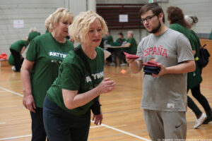 Grupa osób podczas zajęć sportowych w hali gimnastycznej. Widać dwie starsze kobiety o jasnej skórze w zielonych koszulkach z dużym białym napisem „AKTYWNI”; jedna z nich, z krótkimi jasnymi kręconymi włosami, jest na pierwszym planie i jest pochylona do przodu, jakby przygotowywała się do rzutu. Obok stoi młody mężczyzna o jasnej skórze w szarym stroju sportowym z czerwonym napisem „ESPAÑA” na koszulce; ma ciemne włosy, okulary i krótki zarost, i trzyma w dłoniach kilka małych woreczków (czerwone i granatowe), podobnych do woreczków do gry w cornhole. W tle są kolejne osoby w zielonych koszulkach, drewniana podłoga z liniami boiska i małe pomarańczowe znaczniki. W prawym dolnym rogu jest podpis: „fot. Magdalena Seredyńska / Uniwersytet Szczeciński”.