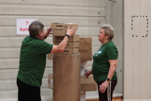 Stos kartonowych pudeł ustawiony pionowo na środku kadru, z kilkoma mniejszymi pudełkami ułożonymi na większym, wysokim kartonie. Po lewej stronie stoi mężczyzna w zielonej koszulce i ciemnych spodniach, z krótkimi szarymi włosami; rękami podtrzymuje i układa górne pudełka. Po prawej stronie stoi kobieta w zielonej koszulce i ciemnych spodniach z jasnym paskiem z boku, z krótkimi jasnoszarymi włosami; ma zegarek na lewym nadgarstku i pierścionek na dłoni. Tło stanowi jasna, metalowa brama lub ściana z poziomymi przetłoczeniami. Po lewej w tle wisi kartka z czerwonym napisem: „7 Kart…” (reszta ucięta). W prawym dolnym rogu widoczny podpis: „fot. Magdalena Seredyńska/Uniwersytet Szczeciński”.
