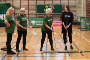 Cztery kobiety na sali gimnastycznej podczas zajęć sportowych z elementami minigolfa. Trzy starsze kobiety stoją po lewej stronie w zielonych koszulkach z napisem „AKTYWNI”; jedna z nich trzyma kij golfowy i stoi przy zielonej macie do puttowania, na której leżą dwie białe piłeczki. Po prawej stoi młodsza kobieta o prostych, ciemnych włosach w czarnej koszulce z napisem „AKTYWNI”, także trzyma kij. Na podłodze widać rozstawione kolorowe pachołki. W tle wiszą banery: zielony z napisem „FUNDACJA LOTTO” oraz tablica z godłem i napisem „Ministerstwo Sportu i Turystyki”. Na dole zdjęcia jest podpis: „fot. Magdalena Seredyńska / Uniwersytet Szczeciński”.
