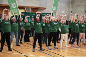 Grupa osób starszych ćwiczących na hali sportowej, stojących w rzędzie z rękami uniesionymi nad głową i splecionymi dłońmi. Większość uczestników ma na sobie zielone koszulki z białymi napisami. Widać kobiety i mężczyzn; część ma siwe lub jasne włosy, niektórzy noszą okulary. Stoją na parkiecie z liniami boiskowymi, w sportowych butach; niektórzy mają spodnie dresowe, inni zwykłe spodnie. W tle są zielone banery oraz elementy hali (ściany, siatka/bramka). W prawym dolnym rogu widnieje mały podpis: „fot. Magdalena Seredyńska/Uniwersytet Szczeciński”.