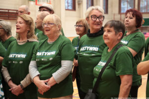 Grupa starszych osób w zielonych koszulkach z nadrukiem „NIEUSTANNIE AKTYWNI” stojąca w hali sportowej. Większość to kobiety o jasnej karnacji; kilka osób ma okulary. Widać różne fryzury: siwe i blond, krótkie i średniej długości; jedna kobieta ma krótkie siwe włosy i duże czarne okulary, inna ma krótkie ciemne włosy, a jeszcze inna ma rude włosy. W tle są kolejne osoby w podobnych koszulkach, fragmenty ścian sali gimnastycznej oraz elementy wyposażenia sportowego. Na dole po prawej jest mały napis: „fot. Magdalena Seredyńska/Uniwersytet Szczeciński”.