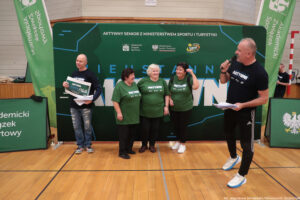 Scena z wydarzenia sportowego w hali, z grupą osób stojących przed dużą zieloną ścianką promocyjną. Na środku stoją trzy starsze kobiety w zielonych koszulkach z napisem „AKTYWNI”; dwie mają ciemne włosy, jedna ma krótkie białe włosy. Stoją blisko siebie i uśmiechają się, jak do pamiątkowego zdjęcia. Po lewej stronie stoi łysy mężczyzna w czarnej koszulce „AKTYWNI”, trzyma duży symboliczny czek i kilka kartek. Na czeku widoczny jest tekst „VOUCHER 250 zł”. Po prawej stronie stoi mężczyzna w czarnej koszulce „AKTYWNI”, w dresowych spodniach, mówi do mikrofonu i trzyma plik kartek.
