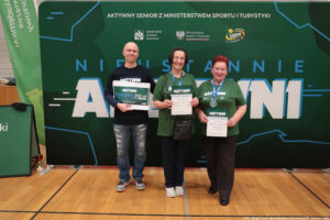 Grupa trzech dorosłych osób stojących w hali sportowej przed dużą zieloną ścianką eventową. Wszyscy stoją w jednym rzędzie i trzymają w dłoniach dyplomy/certyfikaty; osoba skrajnie z lewej trzyma dodatkowo duży bon. Tło to zielona ścianka z napisami i logotypami.