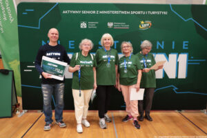 Grupa pięciu dorosłych osób stojących w hali sportowej przed dużą zieloną ścianką eventową. Wszyscy stoją w jednym rzędzie i trzymają w dłoniach dyplomy/certyfikaty; osoba skrajnie z lewej trzyma dodatkowo duży bon. Tło to zielona ścianka z napisami i logotypami