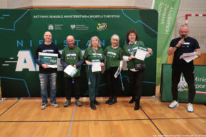 Grupa sześciu dorosłych osób stojących w hali sportowej przed dużą zieloną ścianką promocyjną. Cztery osoby na środku pozują do zdjęcia i trzymają dyplomy lub tablicę z nagrodą, a po prawej stronie mężczyzna w ciemnym stroju mówi do mikrofonu, trzymając kartki.