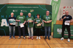 Grupa sześciu dorosłych osób stojących w hali sportowej przed dużą zieloną ścianką promocyjną. Cztery osoby na środku pozują do zdjęcia i trzymają dyplomy lub tablicę z nagrodą, a po prawej stronie mężczyzna w ciemnym stroju mówi do mikrofonu, trzymając kartki.