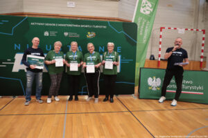 Grupa sześciu dorosłych osób stojących w hali sportowej przed dużą zieloną ścianką promocyjną. Cztery osoby na środku pozują do zdjęcia i trzymają dyplomy lub tablicę z nagrodą, a po prawej stronie mężczyzna w ciemnym stroju mówi do mikrofonu, trzymając kartki.